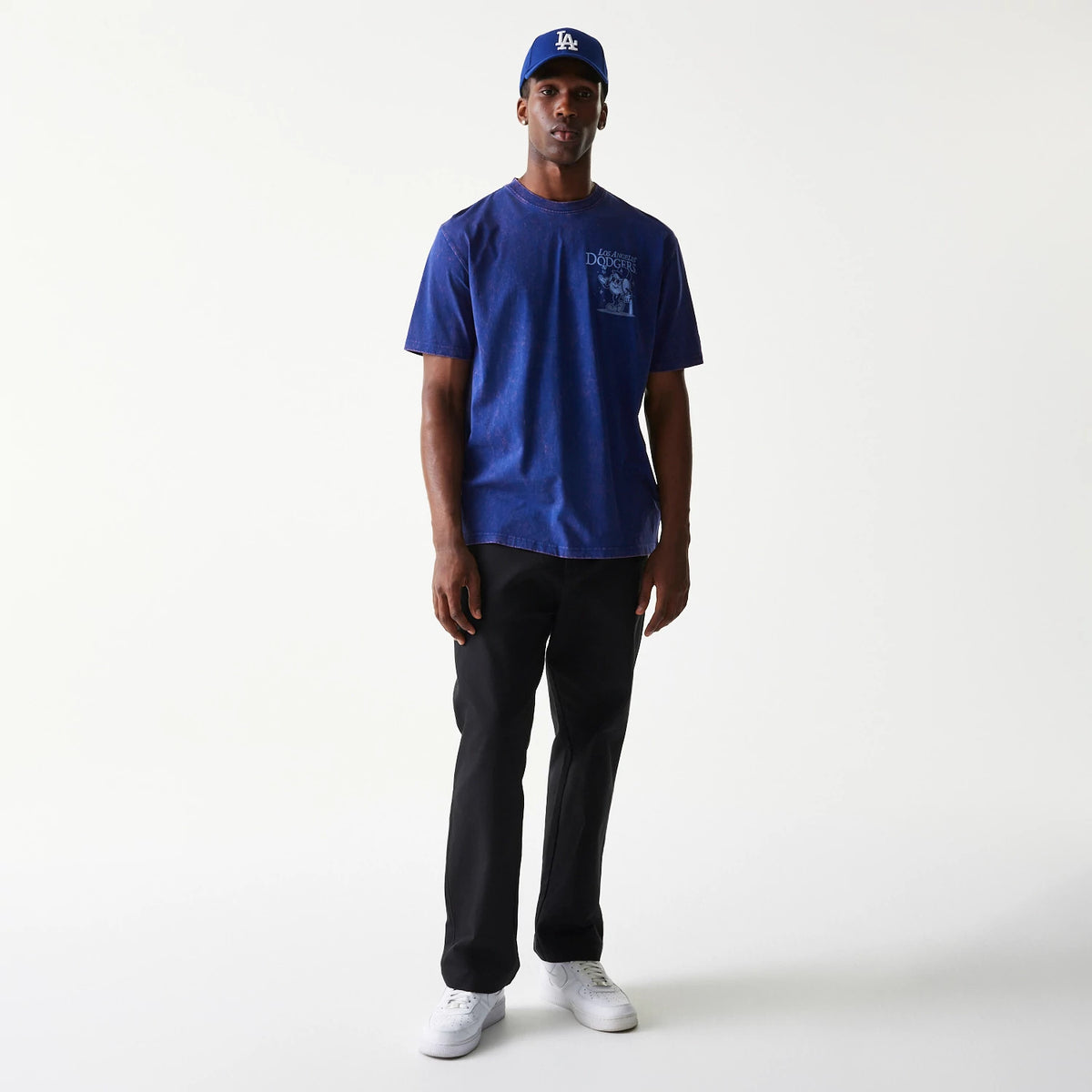 New Era LA Dodgers MLB Character Dark Blue T-Shirt, on-model showing a man wearing a dark blue t-shirt with a Los Angeles Dodgers graphic on the left chest, paired with black trousers and a blue cap featuring a raised LA logo.