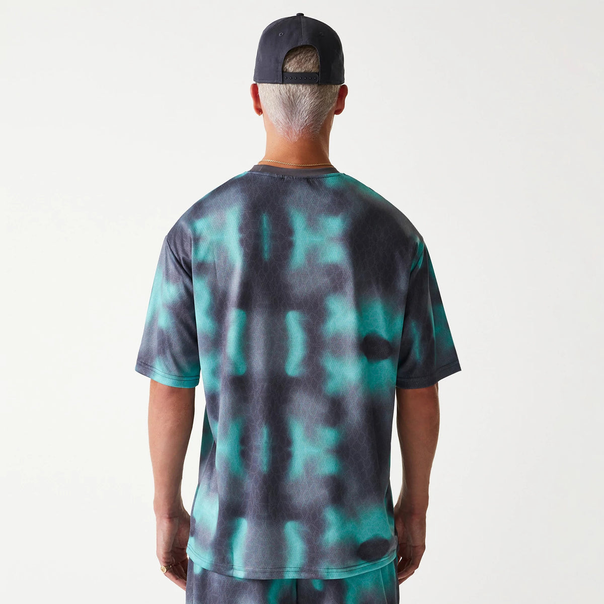 New Era Chicago Bulls NBA All Over Print Green Oversized T-Shirt, rear view showing the green and black tie-dye pattern covering the back, with short sleeves and a relaxed fit, worn by a person with a black cap featuring a row of eyelets on the back.