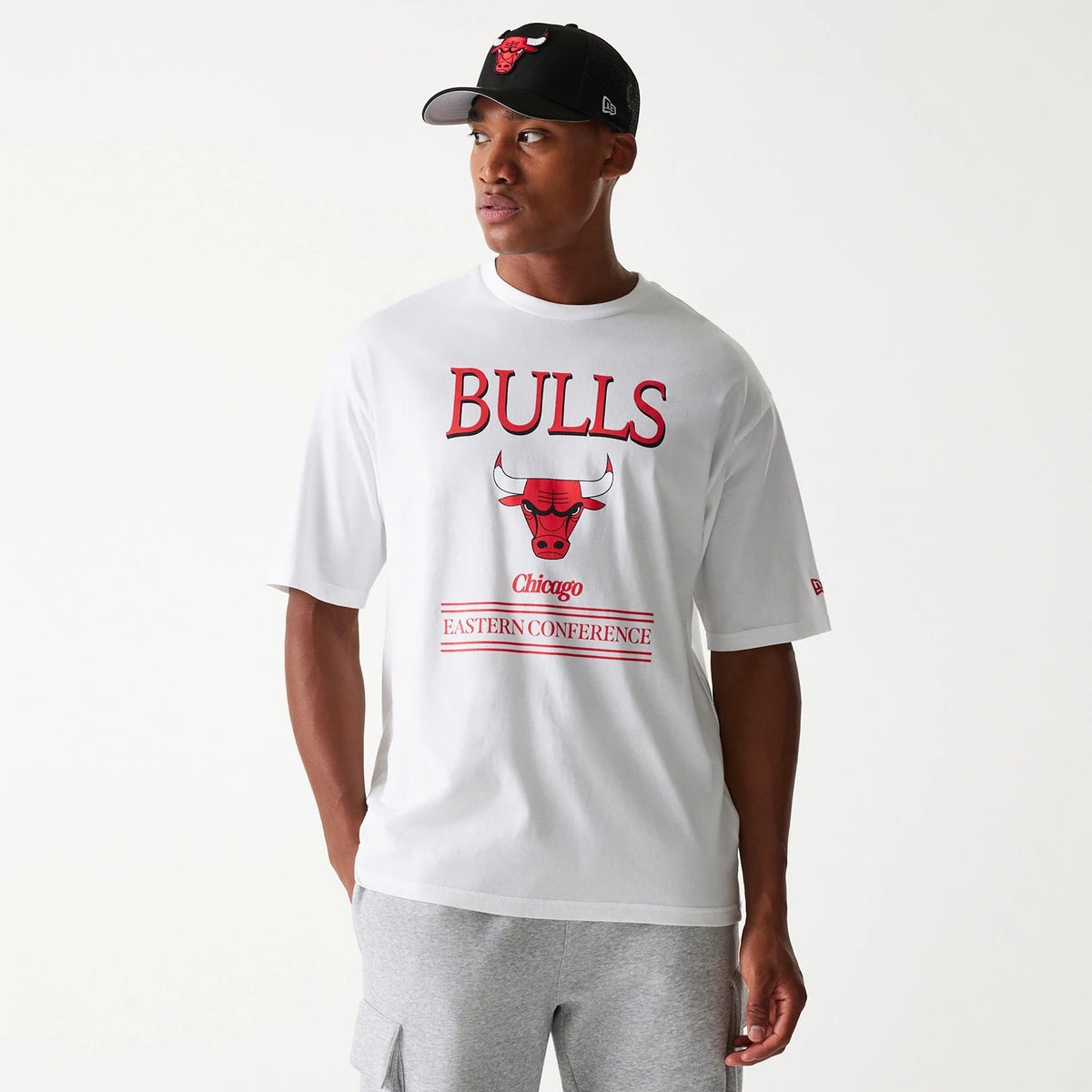 New Era Chicago Bulls NBA Team Graphic White Oversized T-Shirt, on-model view showing the bold red Bulls and Eastern Conference text with a large red bull graphic on the chest, paired with a black New Era cap featuring a raised Bulls logo.
