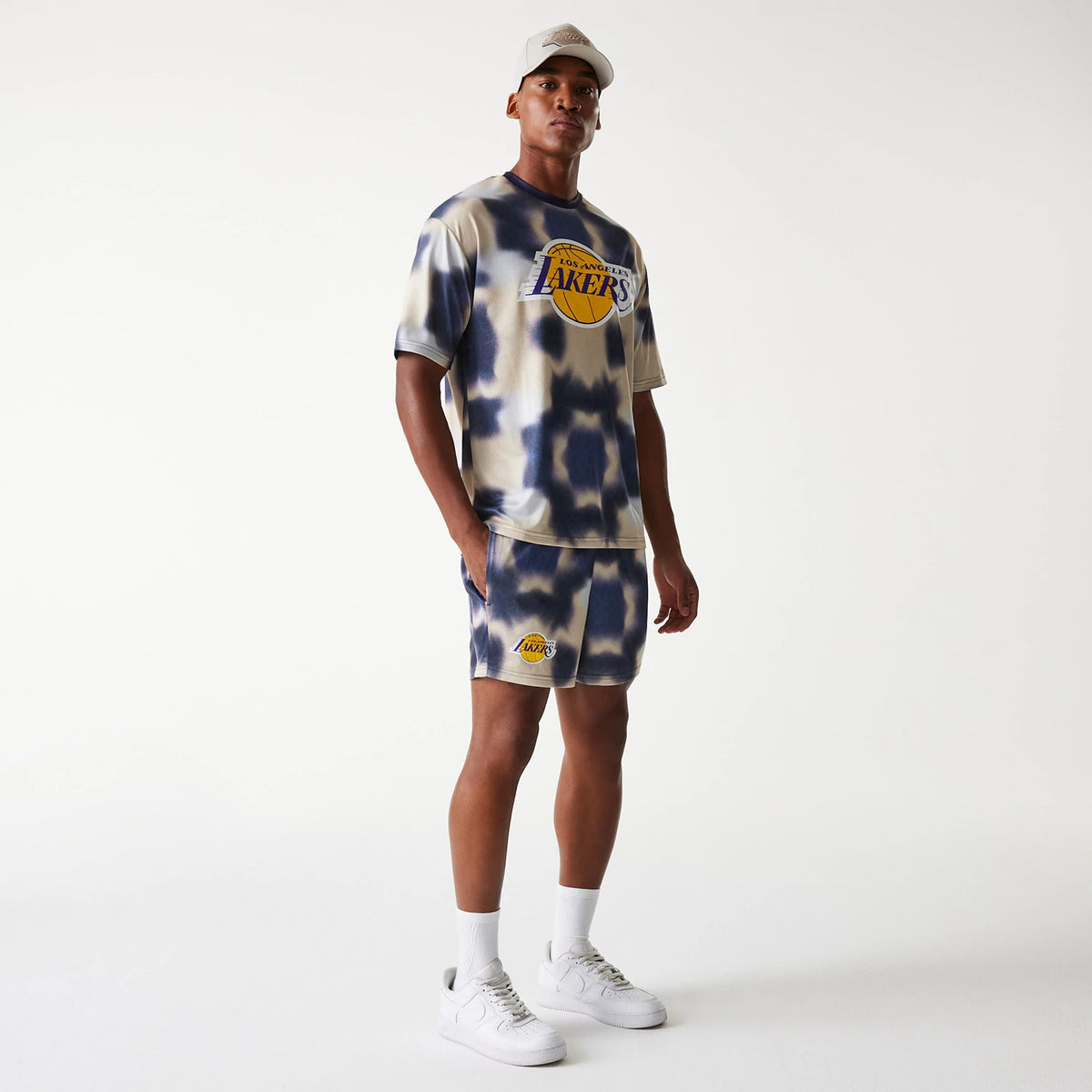 New Era LA Lakers NBA All Over Print Cream Shorts, on-model view showing a person wearing cream shorts with a large printed Lakers logo on the left leg, paired with a matching cream T-shirt and white trainers.