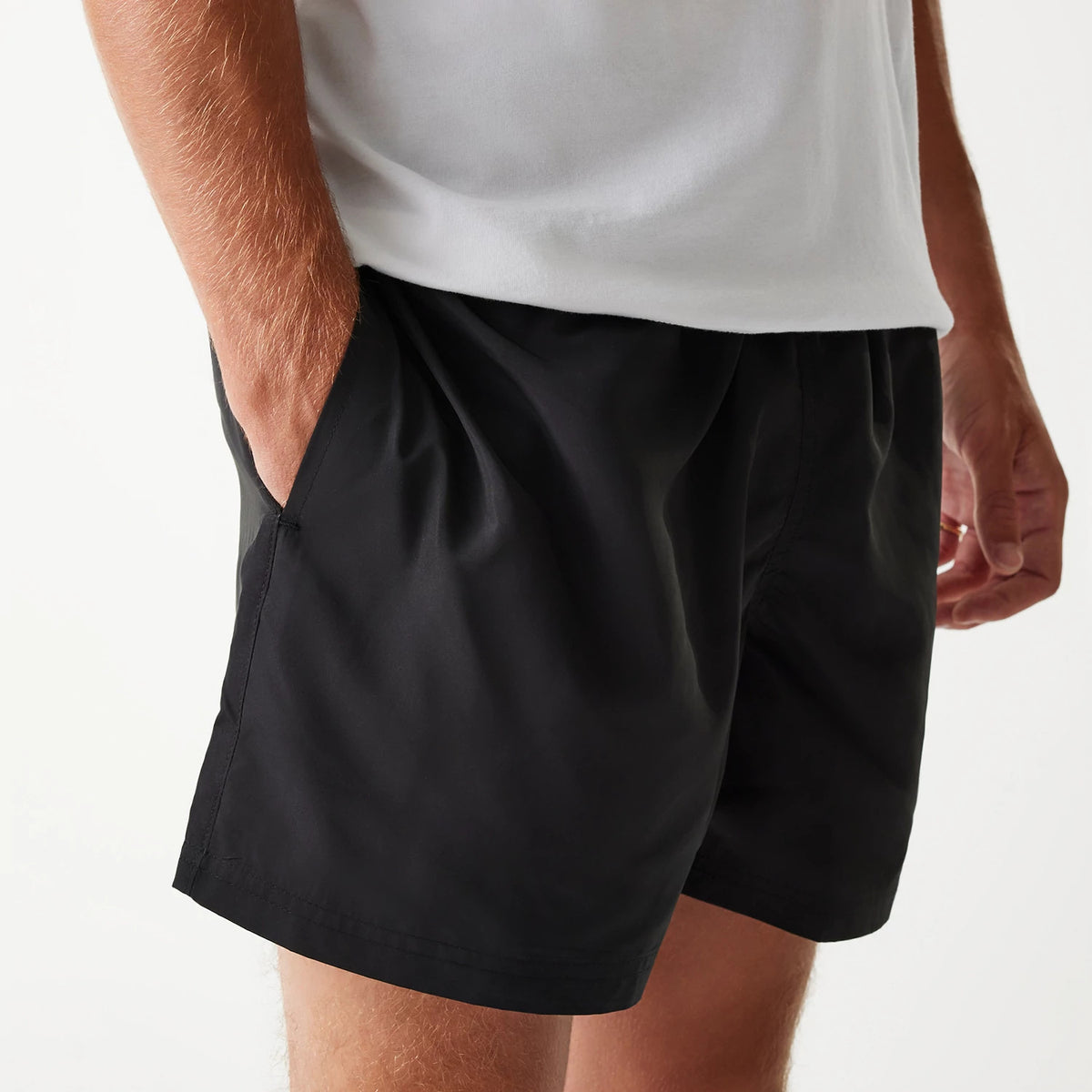 New Era Black Swim Shorts, on-model view showing the relaxed fit black shorts with hands in side pockets, a plain waistband, and the black fabric flowing smoothly down to mid-thigh length.