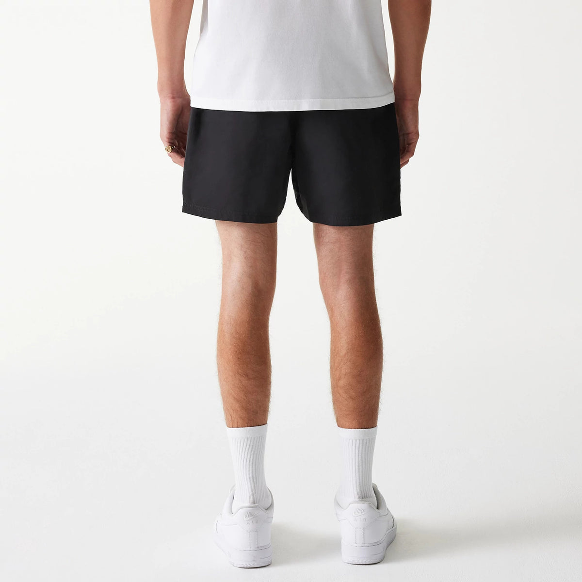 New Era Black Swim Shorts, rear view showing the plain black fabric with a straight hem resting above the knee, worn by a person in a white t-shirt and trainers, contrasting the simplicity of the design.