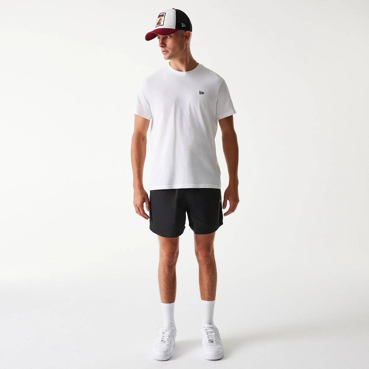 New Era Black Swim Shorts, on-model view showing the black swim shorts with an elasticated waistband paired with a white t-shirt featuring a small raised New Era logo on the chest, and a white and burgundy cap.