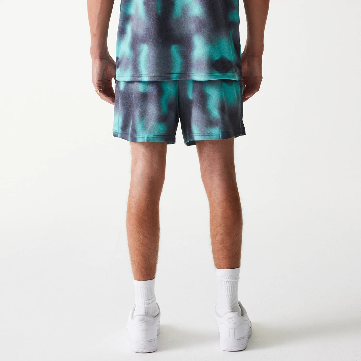 New Era Chicago Bulls NBA All Over Print Green Shorts, rear view showing a blend of green and black patterns on loose-fitting shorts, paired with the matching top worn by a model standing with white sneakers and socks.