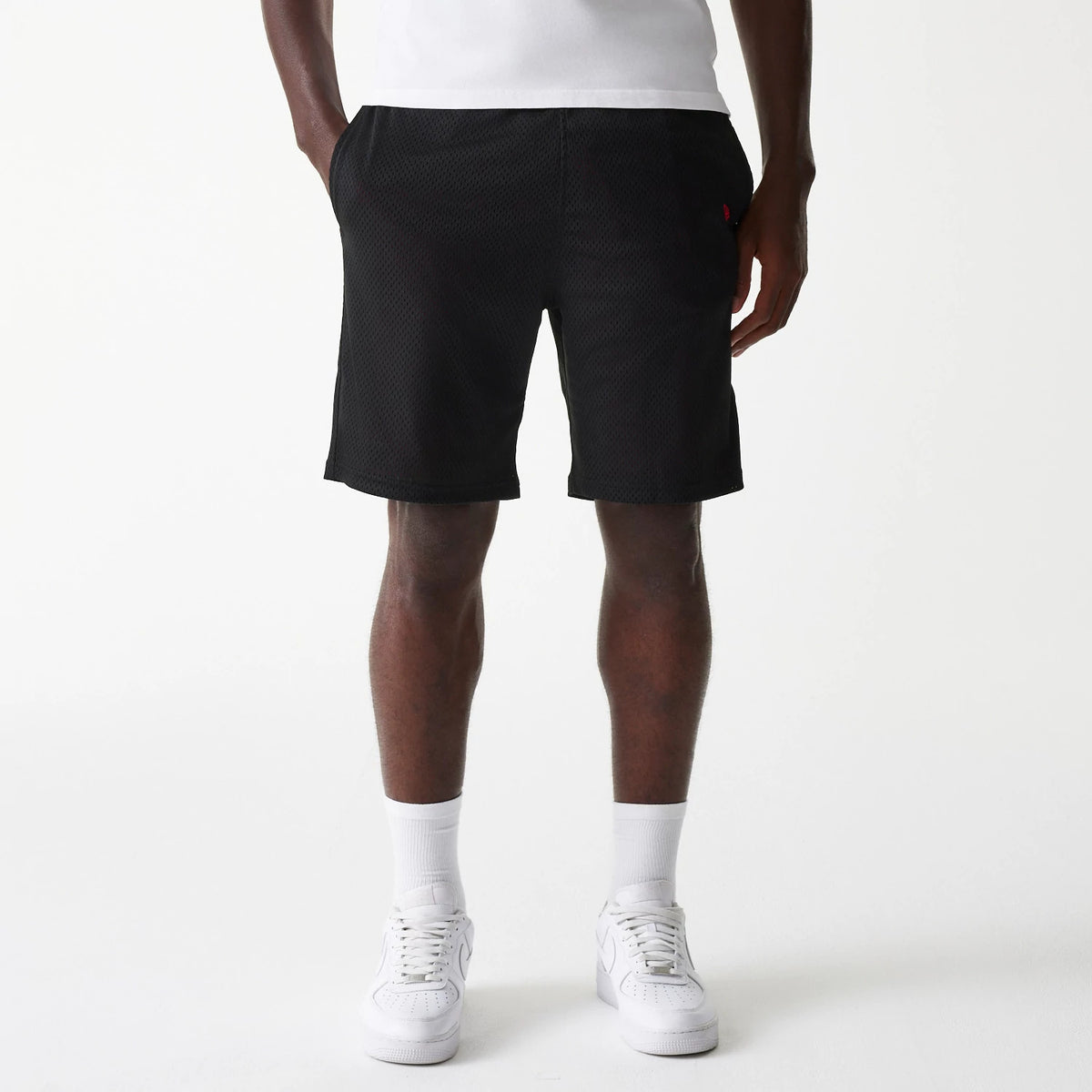 New Era Black Mesh Shorts, front view showing the relaxed fit black mesh fabric with an elastic waistband and side pockets, featuring a small embroidered red logo on the left side near the pocket.