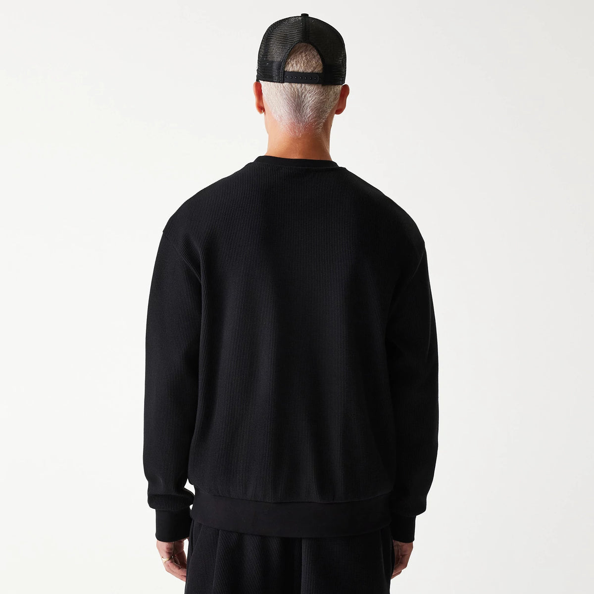 New Era Waffle Black Oversized Crew Neck Sweater, back view on a model wearing matching black shorts and a black cap, showing the sweater's textured waffle knit and relaxed fit with ribbed cuffs and hem.