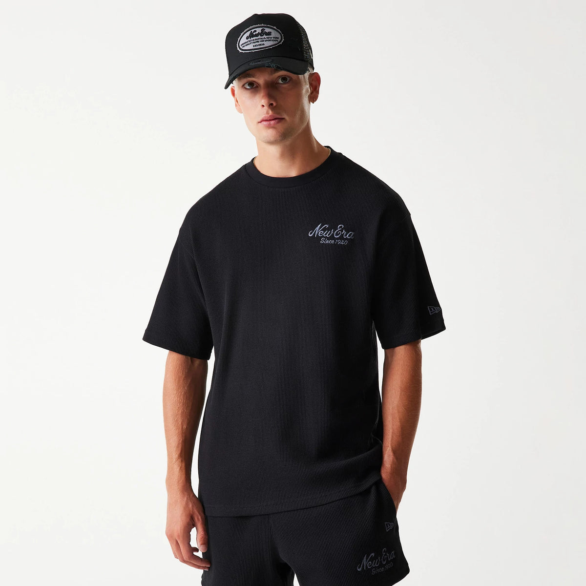 New Era Waffle Black Oversized T-Shirt, on-model view showing the black textured fabric with a white embroidered New Era logo on the left chest and sleeve, paired with a black cap featuring a raised patch logo on the front.