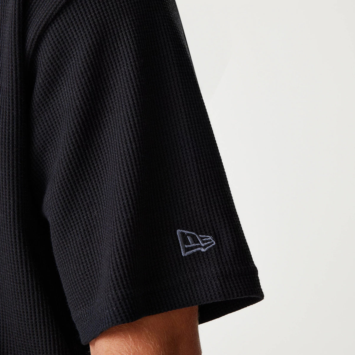 New Era Waffle Black Oversized T-Shirt, side view showing the textured black fabric with a raised New Era logo embroidered in white near the edge of the sleeve.