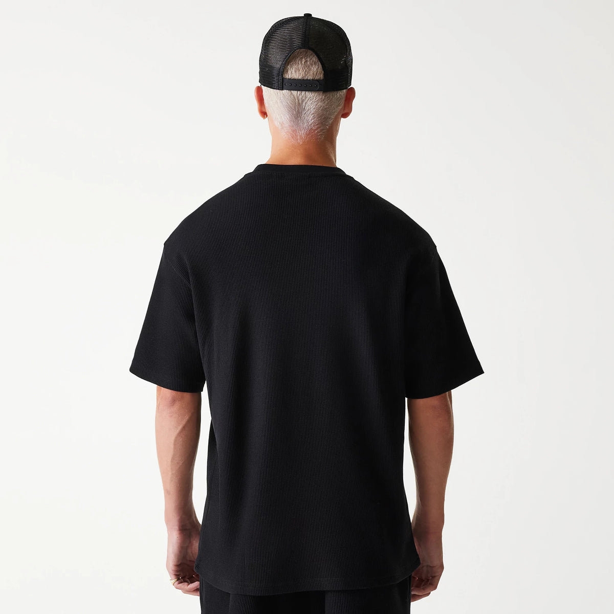 New Era Waffle Black Oversized T-Shirt, rear view showing the solid black back with short sleeves, worn by a person with a mesh black cap featuring a debossed logo, highlighting the relaxed fit of the T-shirt.