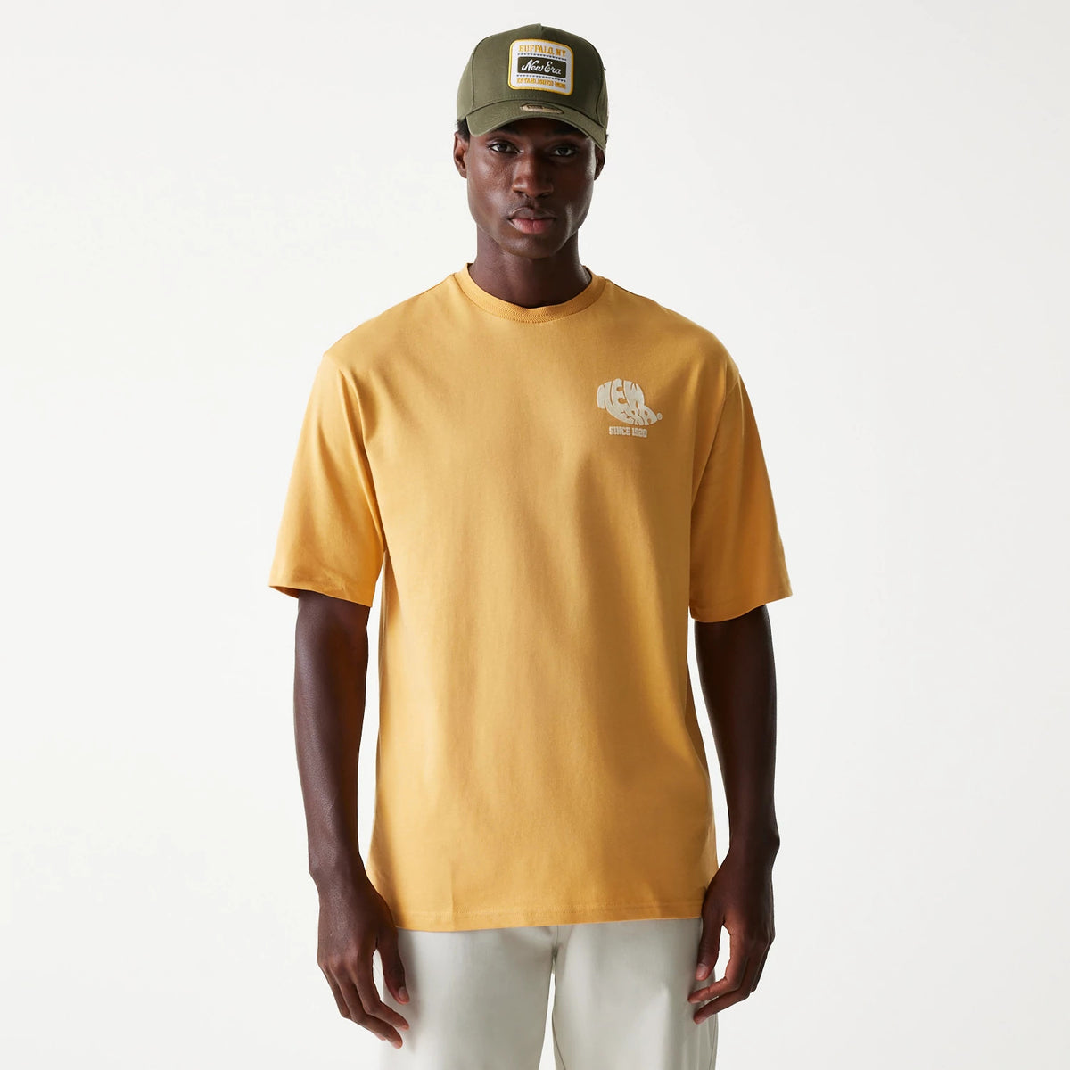 New Era Graphic Beige Oversized T-Shirt, front view showing the beige oversized design with a small embossed graphic logo on the left chest, worn by a model paired with a green cap featuring an embroidered logo on the front panel.