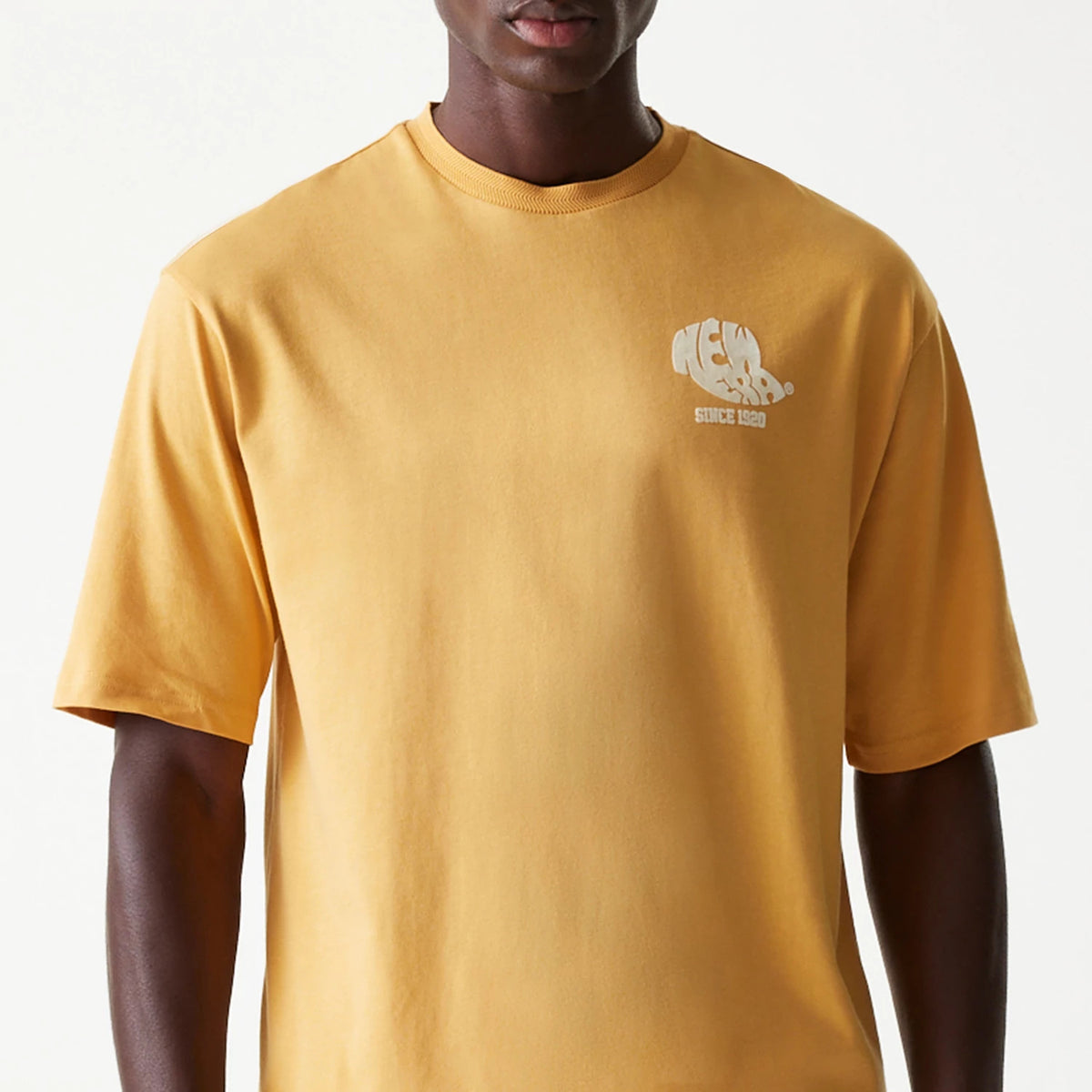 New Era Graphic Beige Oversized T-Shirt, front view showing the short-sleeve beige t-shirt with a white raised New Era logo on the left chest, featuring a relaxed fit and crew neckline.