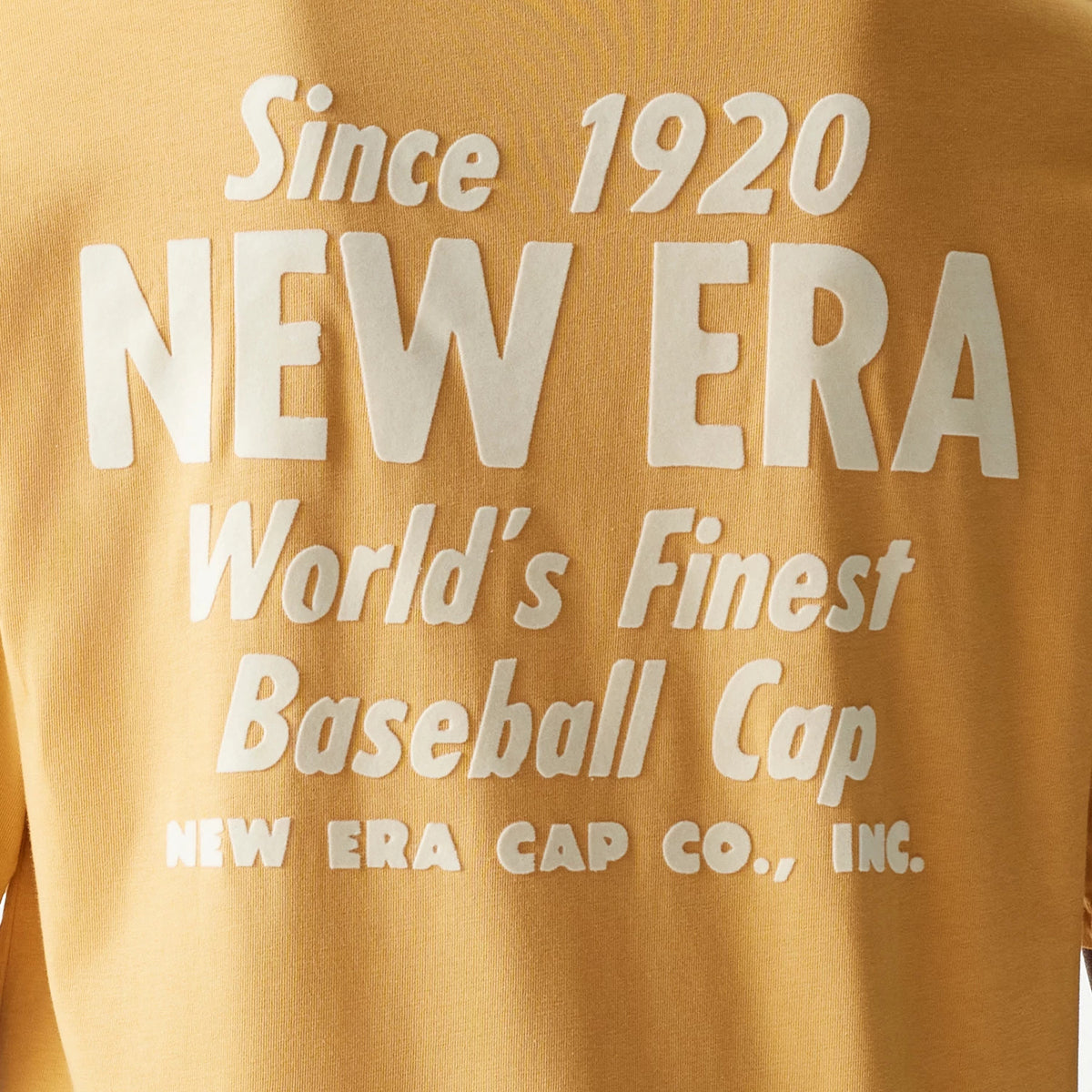 New Era Graphic Beige Oversized T-Shirt, rear view showing large white raised lettering stating Since 1920 NEW ERA World's Finest Baseball Cap NEW ERA CAP CO., INC. on a beige fabric background.