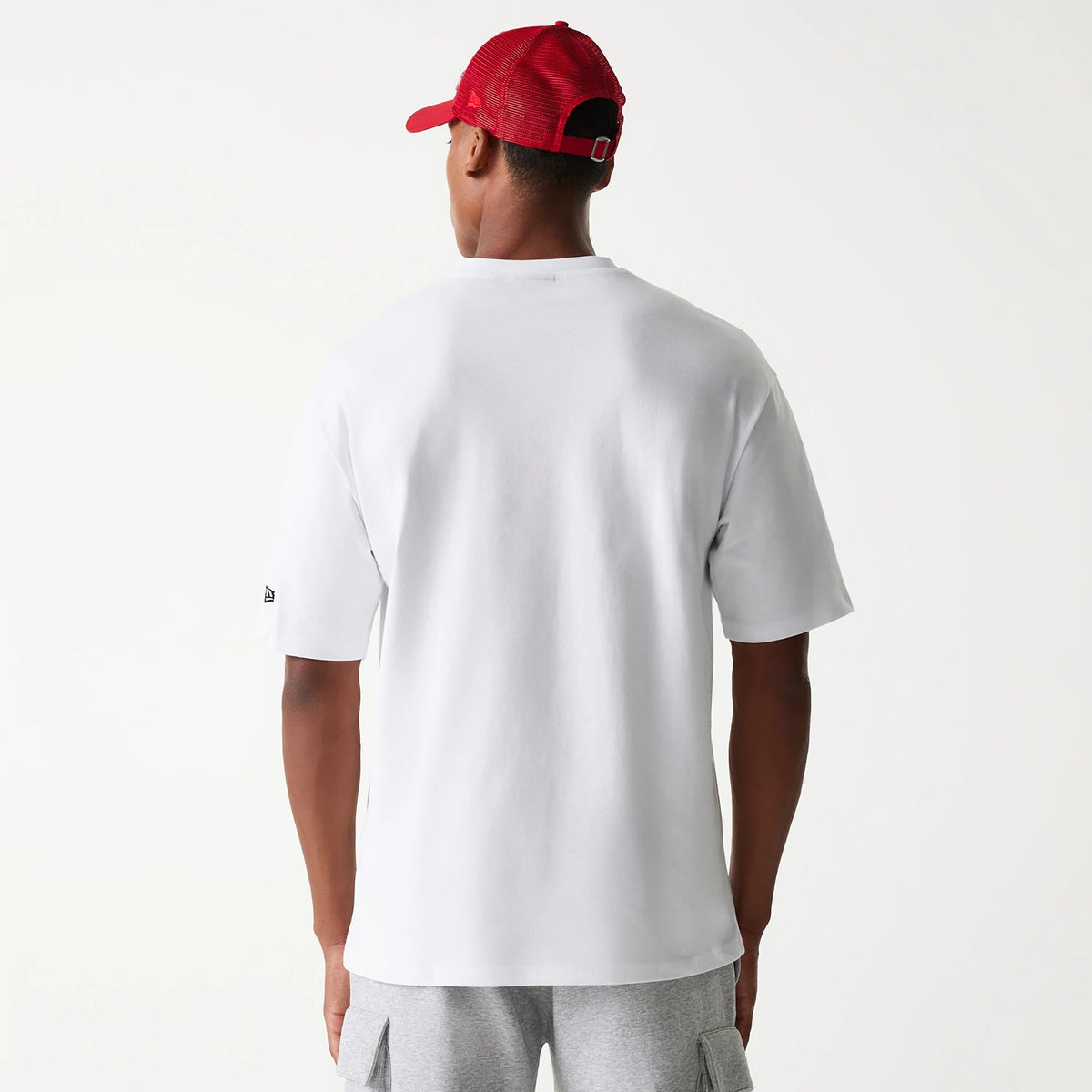 New Era Outdoor Graphic White Oversized T-Shirt, on-model rear view showing the plain white back with a small embroidered New Era logo on the left sleeve, worn with grey shorts and a red cap.