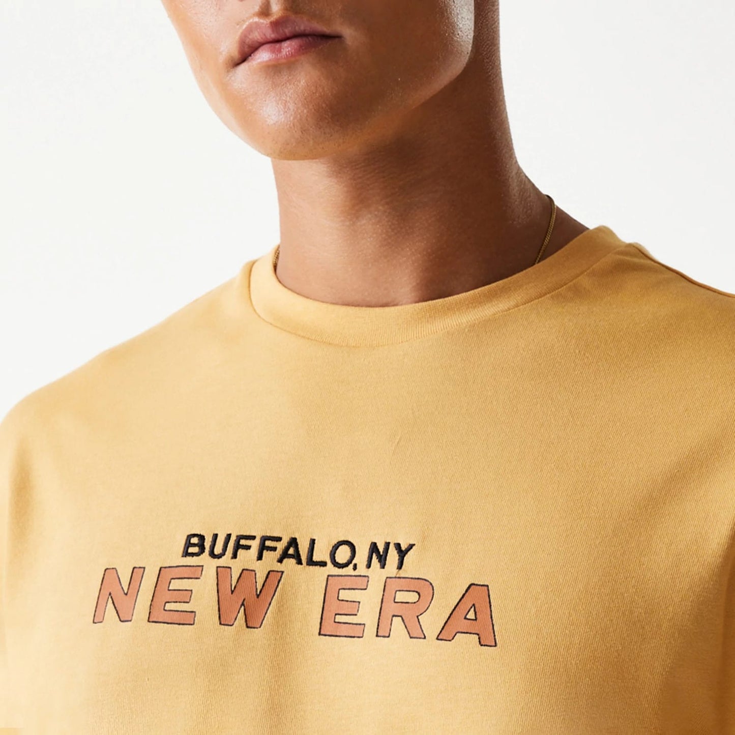 New Era Vacation Graphic Beige Oversized T-Shirt, front view showing the beige fabric with a printed BUFFALO, NY NEW ERA graphic in black and orange letters across the chest, on-model.