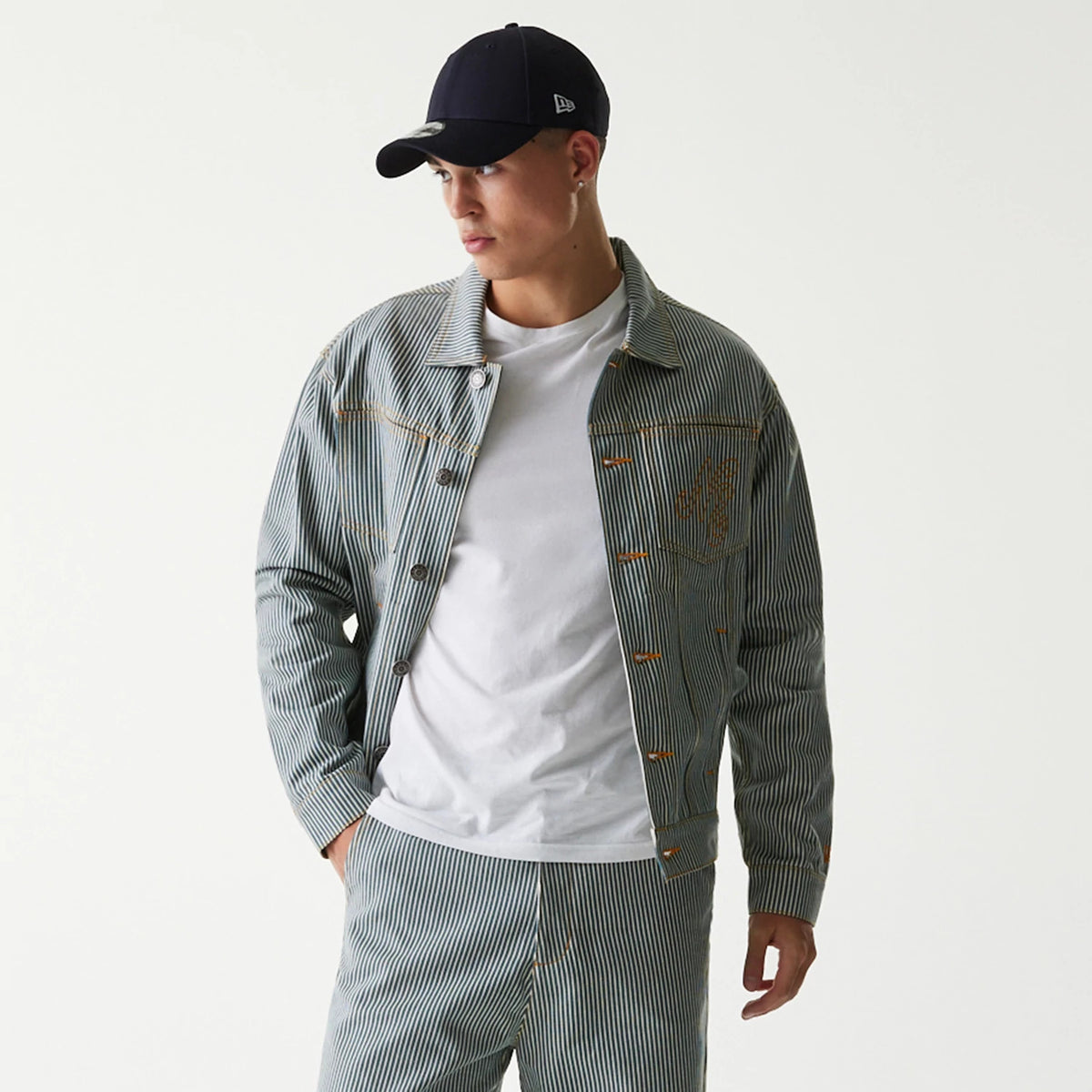 New Era Blue Denim Jacket, on-model view showing a pinstripe pattern with orange stitching detail on the pocket and buttons, worn over a white t-shirt, paired with matching trousers and a navy cap featuring a raised logo on the side.