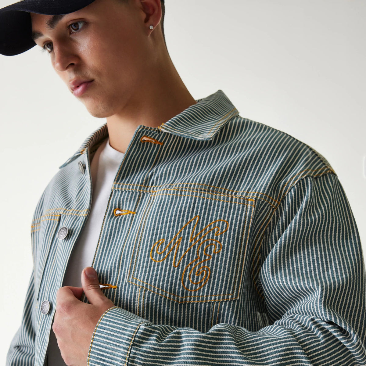 New Era Blue Denim Jacket, on-model view showing a striped denim pattern with orange stitching, a raised New Era logo in orange on the chest pocket and button-up design with contrasting orange buttonholes.