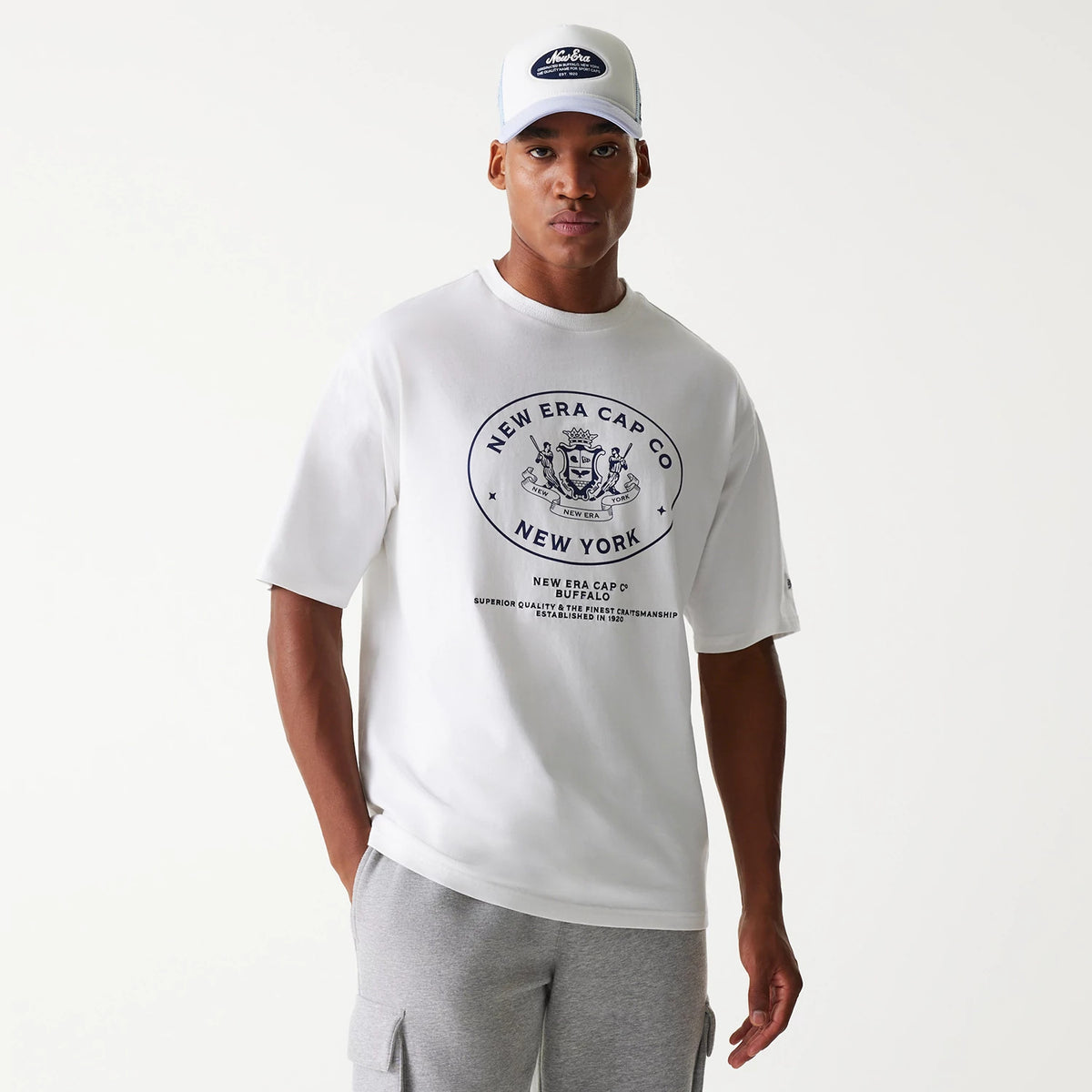New Era Crest White Oversized T-Shirt, on-model view showing the white shirt with a large black crest logo centred on the chest, short sleeves, and a relaxed fit, paired with a white cap featuring a debossed black patch logo.