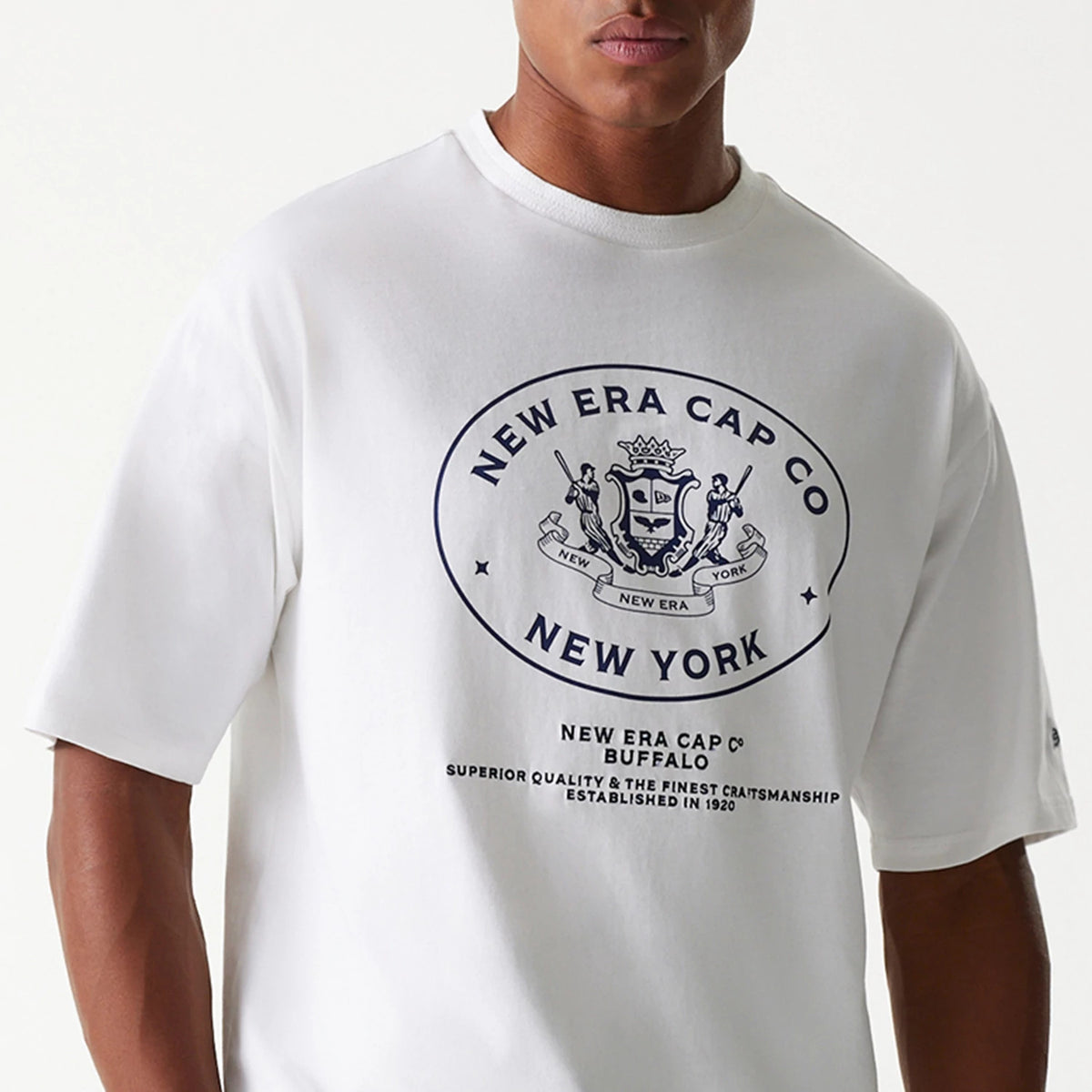 New Era Crest White Oversized T-Shirt, front view showing a large navy crest logo centred on the chest with text beneath, set on a white fabric background, short sleeves and a round neckline.