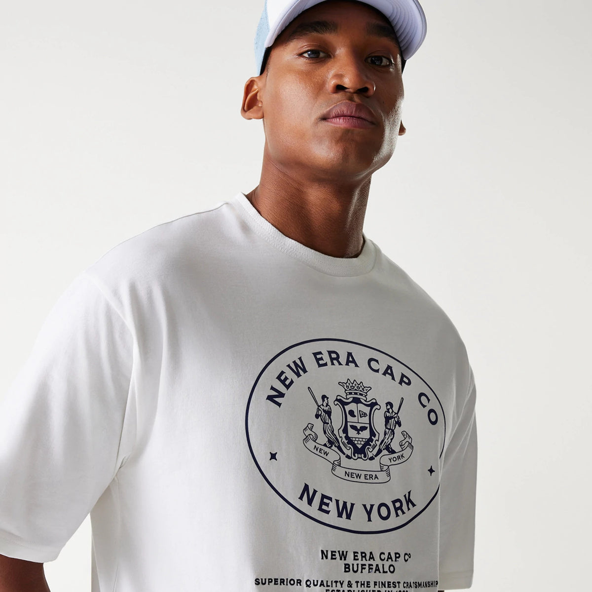 New Era Crest White Oversized T-Shirt, on-model view featuring a large printed crest logo in black on the front, showing a shield surrounded by New Era Cap Co text above New York, worn by a person in a light blue cap.