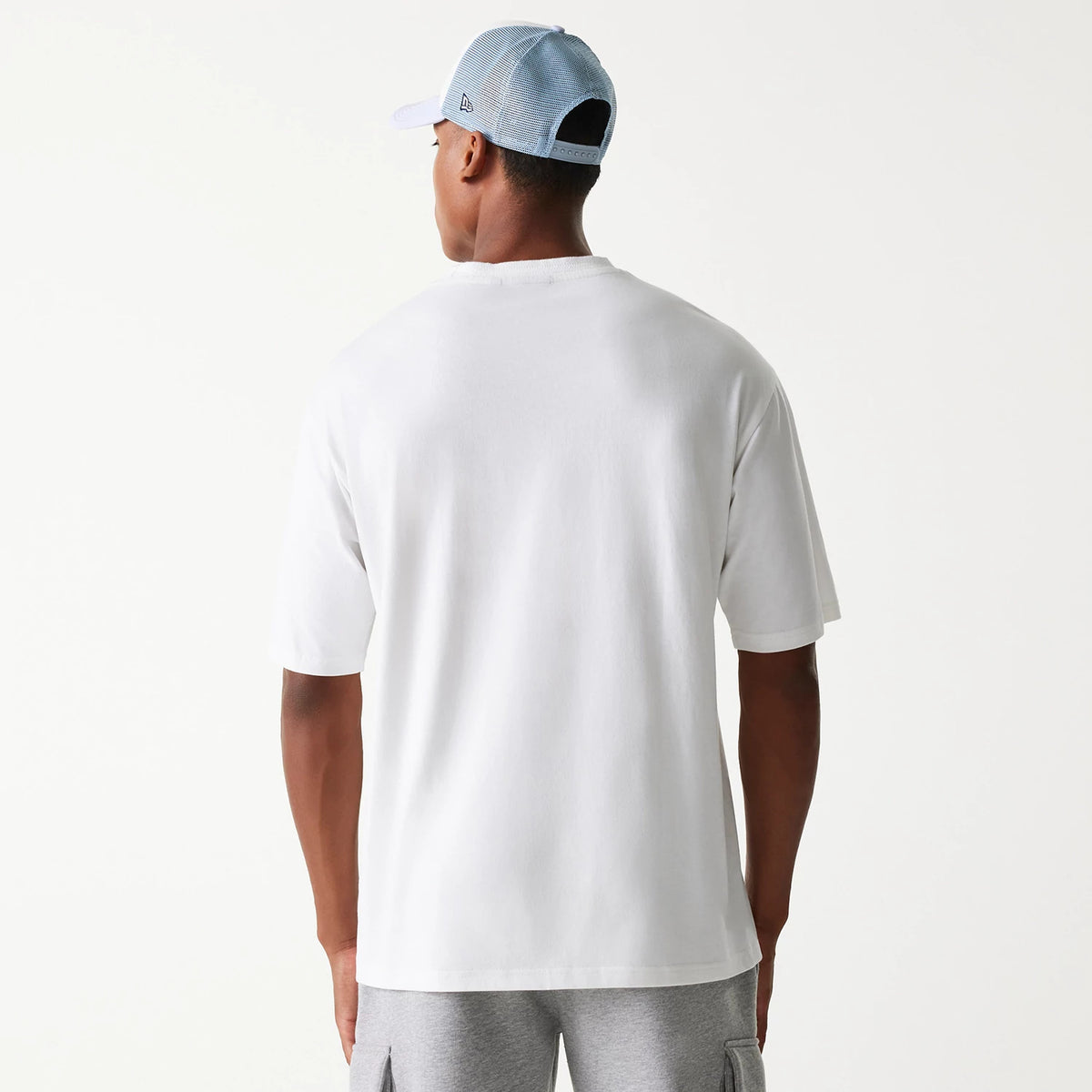 New Era Crest White Oversized T-Shirt, rear view showing the plain white fabric extending down the back, with a relaxed fit across the shoulders and short sleeves, worn with a blue mesh cap featuring a raised New Era logo on the side.