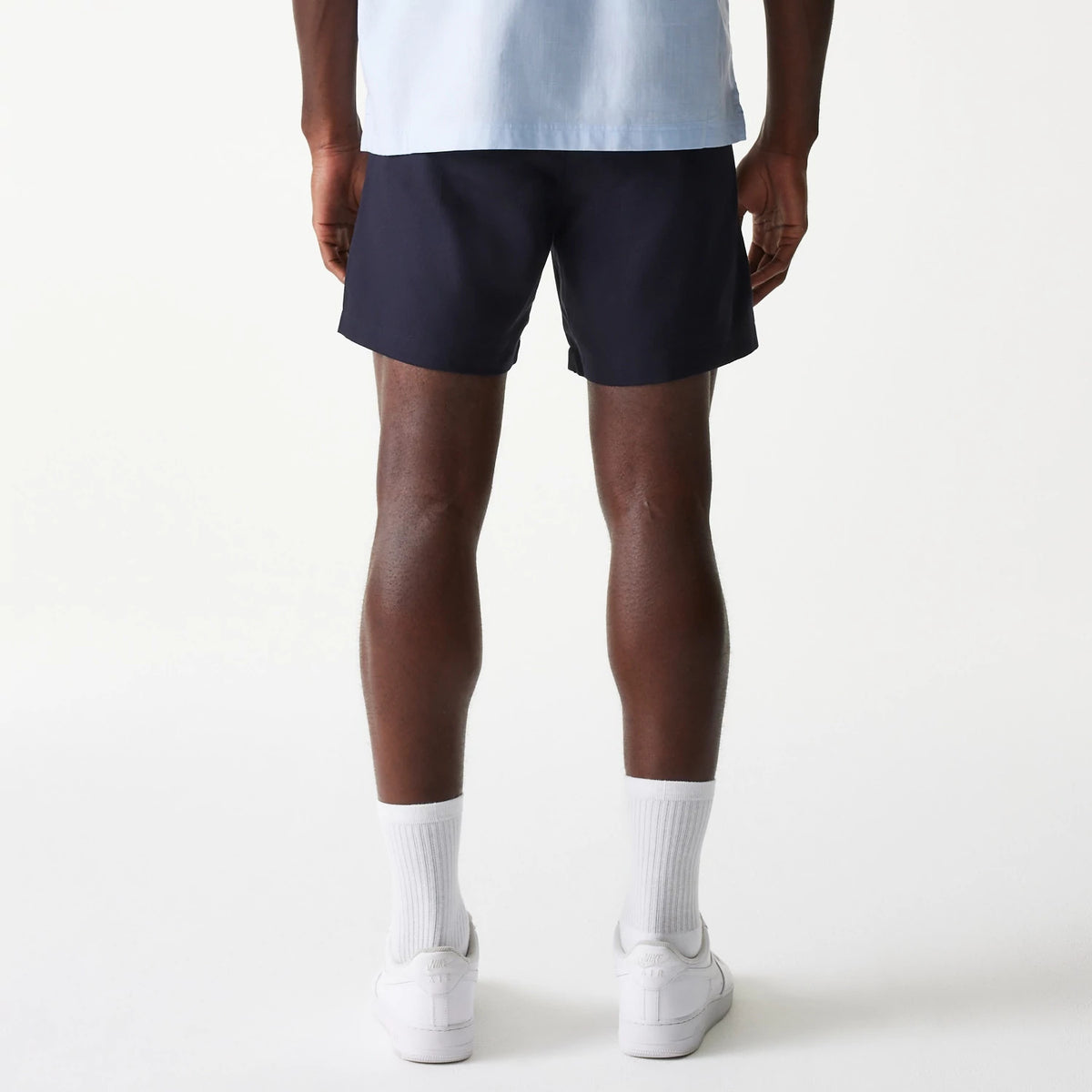New Era Navy Chino Shorts, on-model rear view showing the dark navy shorts with a straight hemline above the knee worn over white socks and white trainers, the plain back complemented by a light blue shirt.