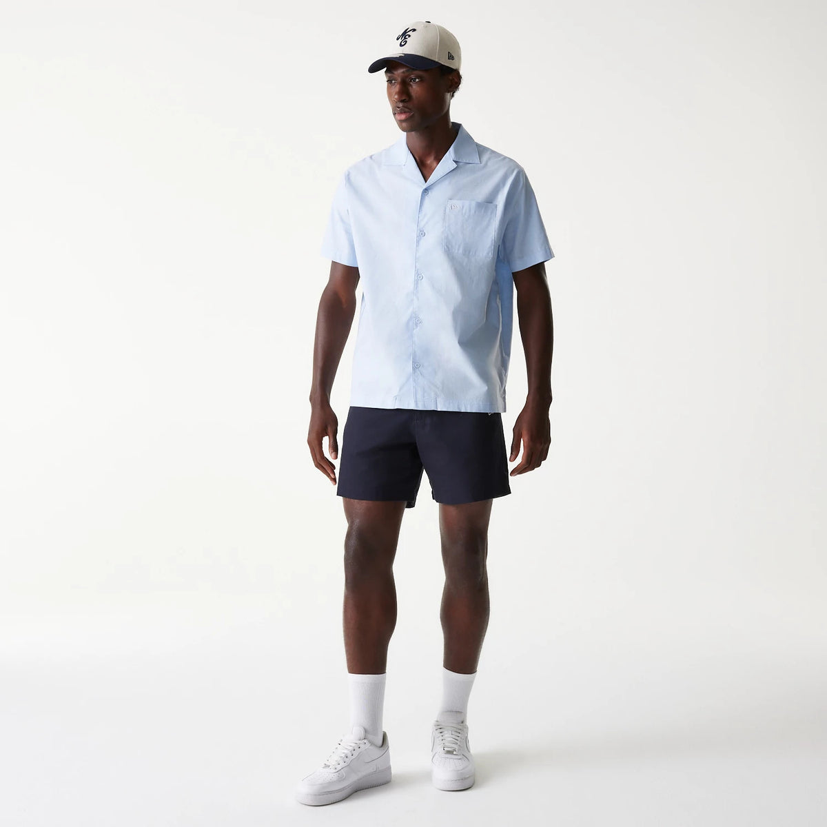 New Era Navy Chino Shorts, on-model view showing the dark navy shorts worn with a light blue shirt and white trainers, a debossed New Era logo on the front of a white cap with a black visor worn by the model.