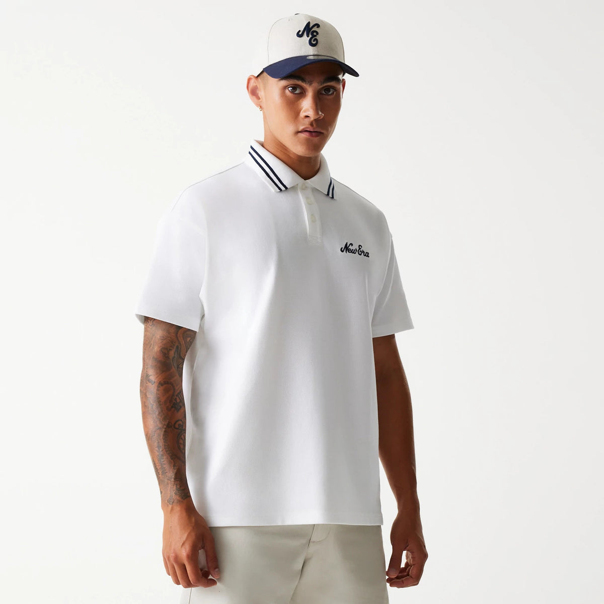 New Era Drop Shoulder White Polo Shirt, front view on a model with an embroidered New Era logo on the left chest, contrasting striped collar, short sleeves, and a model wearing a cap with an embroidered front logo.