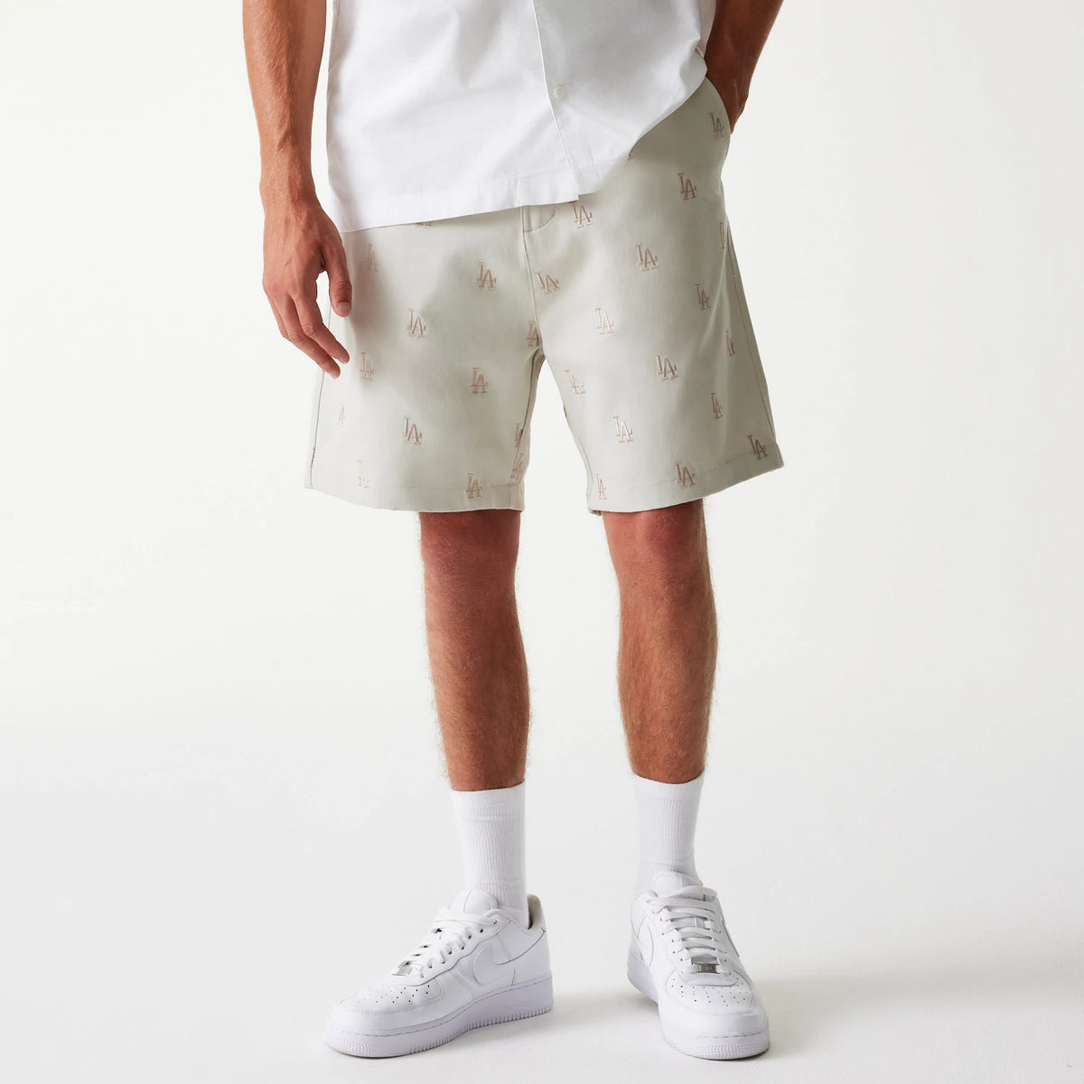 New Era LA Dodgers MLB Monogram Cream Shorts, front view showing cream shorts with repeated debossed LA logos, worn with white socks and shoes, part of a casual outfit with a white shirt.