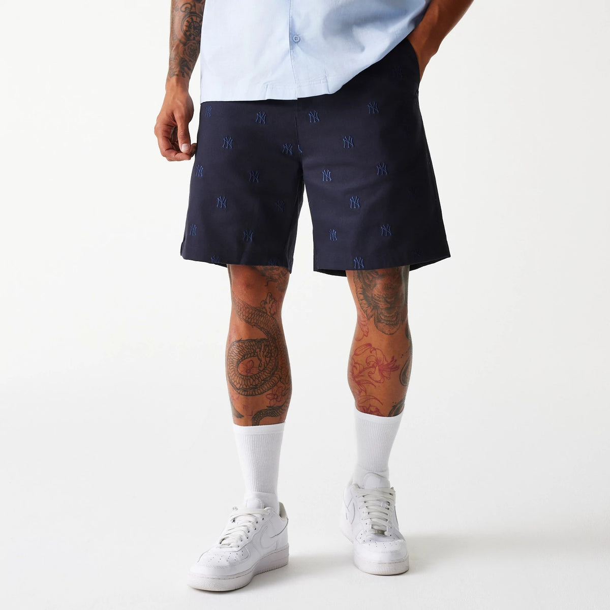 New Era New York Yankees MLB Monogram Navy Shorts, front view showing navy fabric with a repeated embroidered New York Yankees logo pattern in blue and worn by a model with hands in pockets.