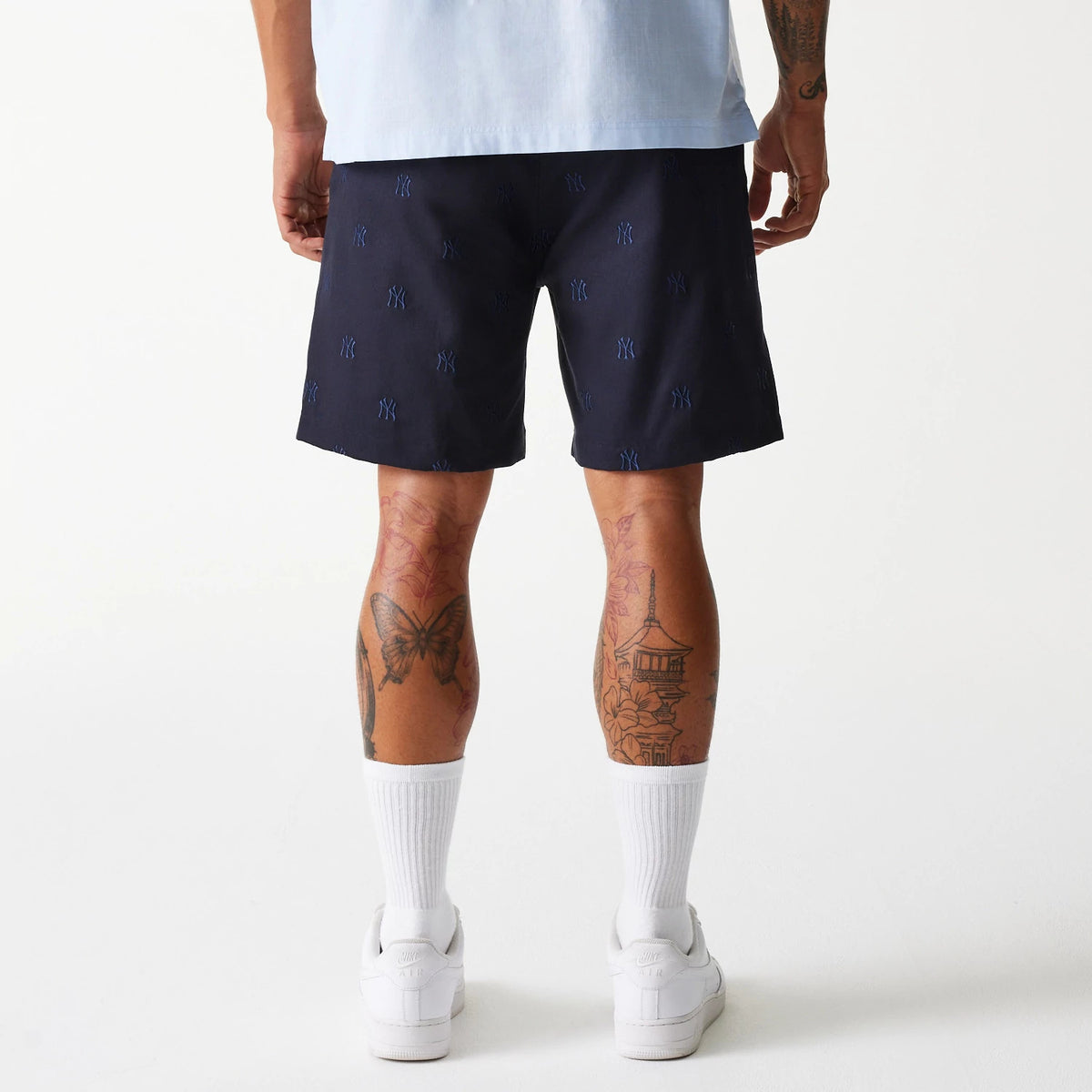 New Era New York Yankees MLB Monogram Navy Shorts, rear view showing navy blue shorts with an all-over raised New York Yankees logo pattern, worn by a tattooed individual standing with white sneakers and socks.