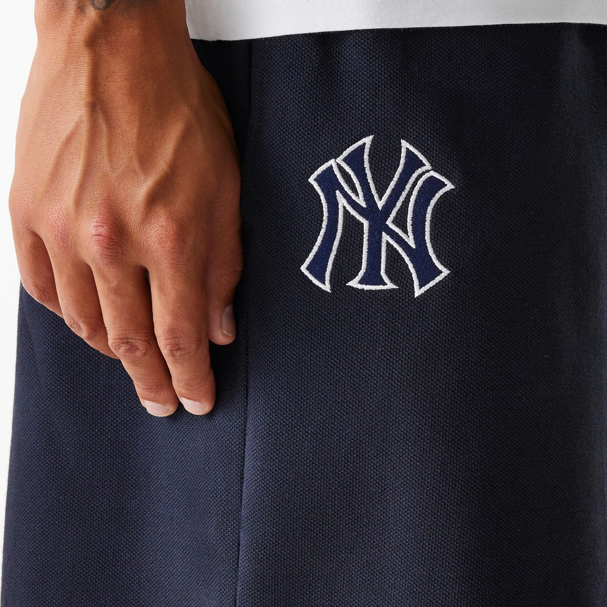 New Era New York Yankees MLB Pique Navy Shorts, side view showing the embroidered Yankees logo in white on the dark navy fabric, with part of a person's hand resting near the logo.