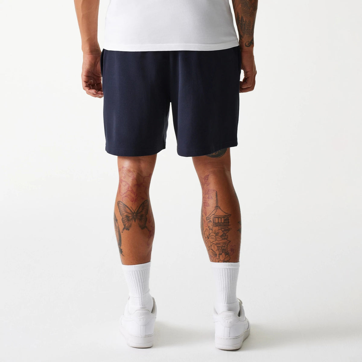 New Era New York Yankees MLB Pique Navy Shorts, rear view showing the plain navy fabric with a straight hem finishing above the knee, worn by a model with tattooed legs, a white shirt, white socks, and white trainers.