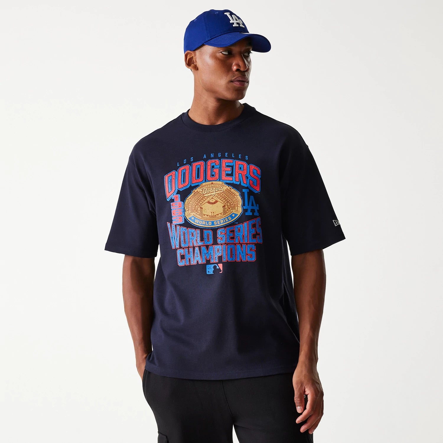 New Era LA Dodgers MLB World Series Navy Oversized T-Shirt, on-model view showing the large front graphic in blue, red, and gold with World Series Champions text, small debossed New Era logo on the sleeve, paired with a blue LA cap.