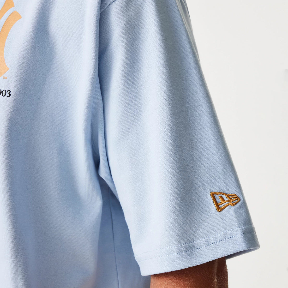 New Era New York Yankees MLB Baseball Graphic Pastel Blue Oversized T-Shirt, close-up view showing the pastel blue sleeve with a raised New Era logo embroidered in gold near the hem, part of the larger graphic partially visible on the chest.