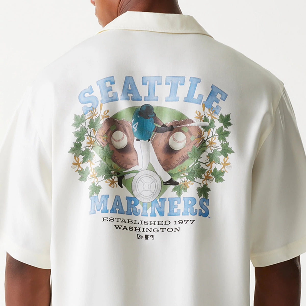 New Era Seattle Mariners MLB Graphic Open White Revere Short Sleeve Shirt, back view on a model showing a large graphic with a baseball player, gloves, and foliage, along with Seattle Mariners text and Established 1977 Washington below.