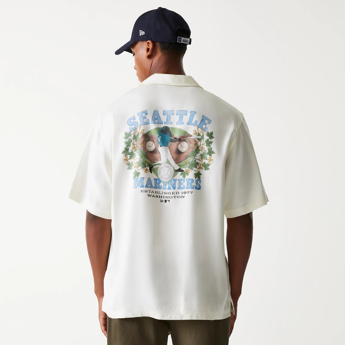 New Era Seattle Mariners MLB Graphic Open White Revere Short Sleeve Shirt, back view on a model wearing a navy cap, highlighting a large graphic with Seattle Mariners text and a baseball player surrounded by foliage and baseball stitching.
