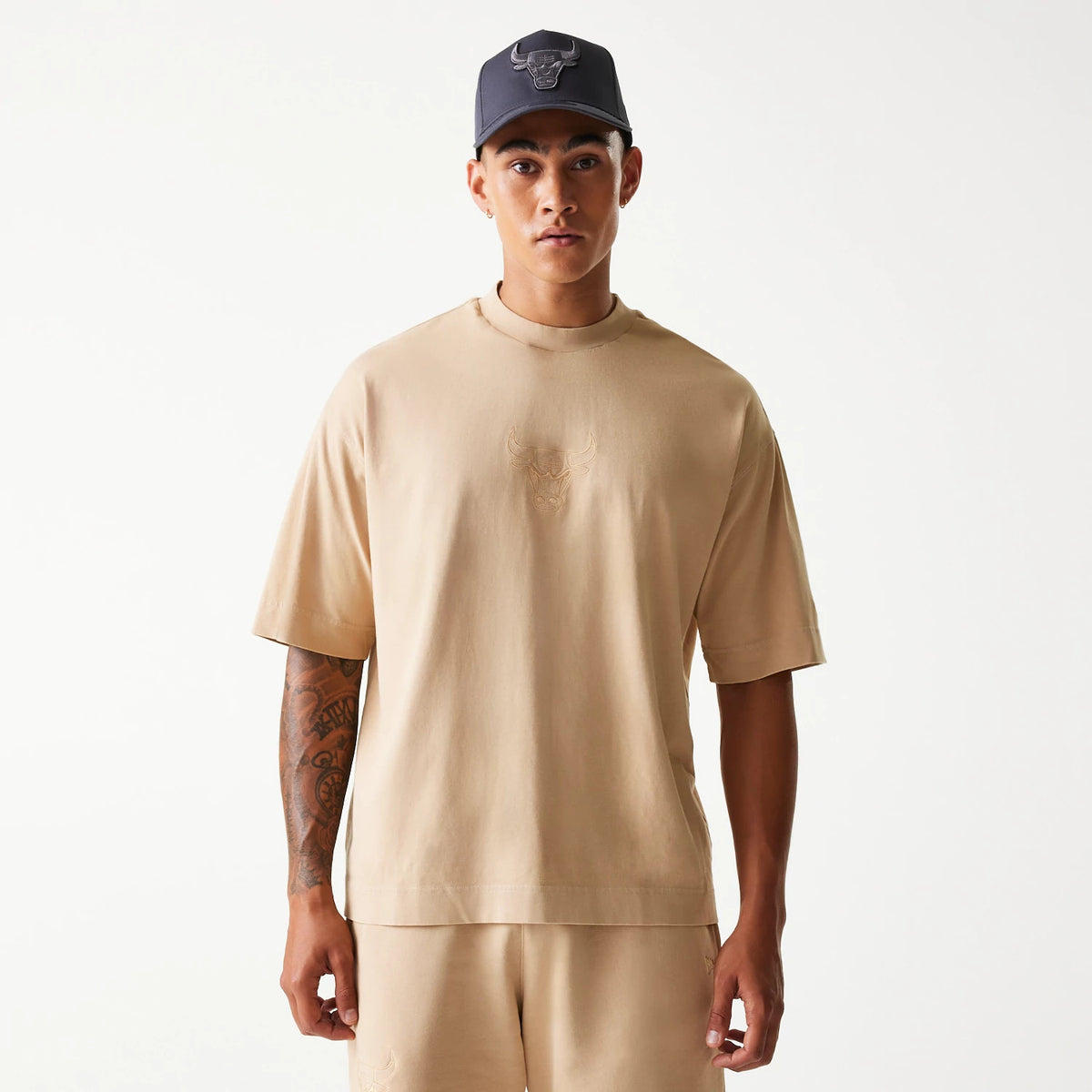 New Era Chicago Bulls NBA Washed Beige Box T-Shirt, front view showing a beige T-shirt with an embossed Chicago Bulls logo centred on the chest worn by a person, paired with beige shorts and a dark cap featuring a raised Bulls logo.