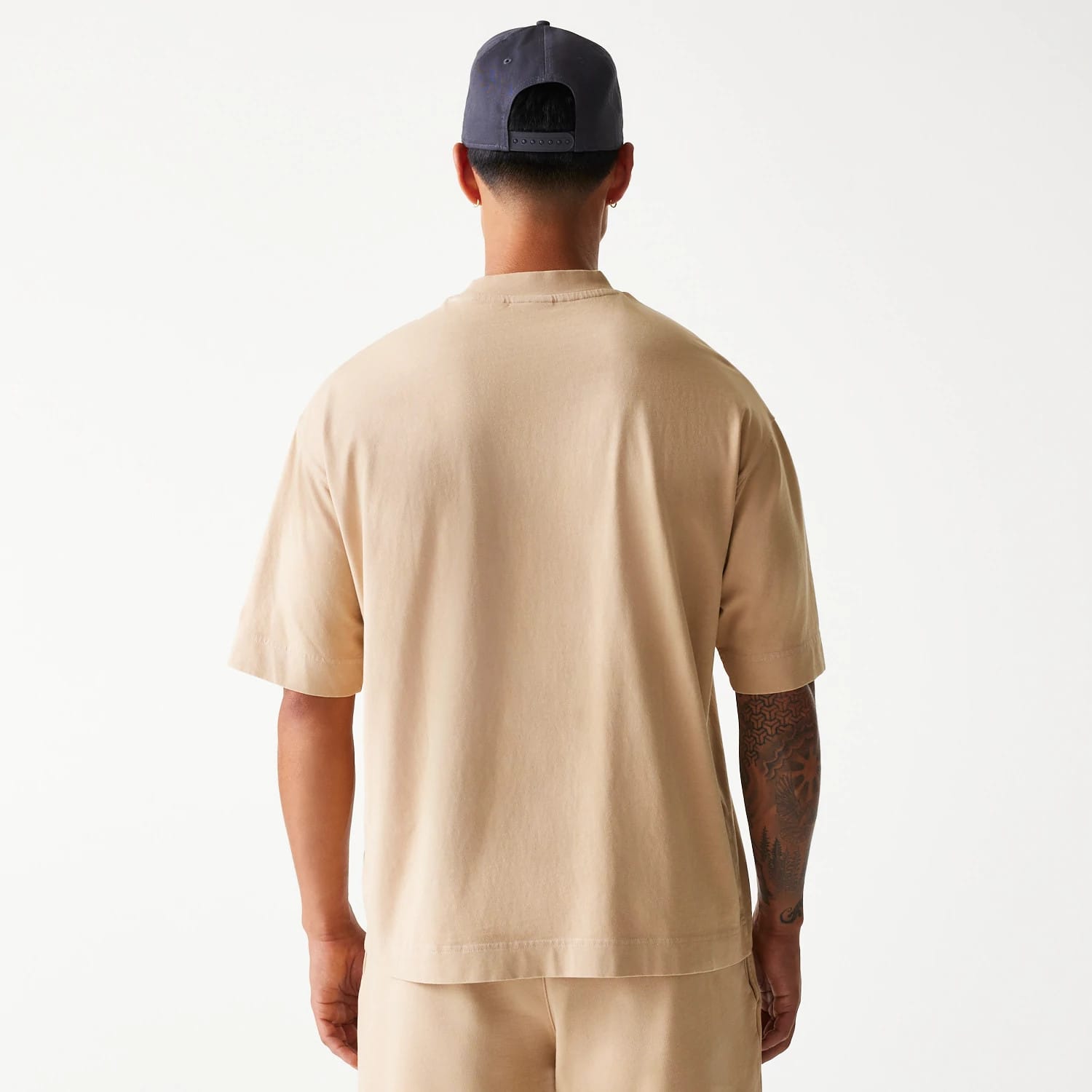 New Era Chicago Bulls NBA Washed Beige Box T-Shirt, rear view showing the plain beige fabric with short sleeves, worn by a model paired with light beige shorts, and a dark cap featuring eyelets worn backwards.
