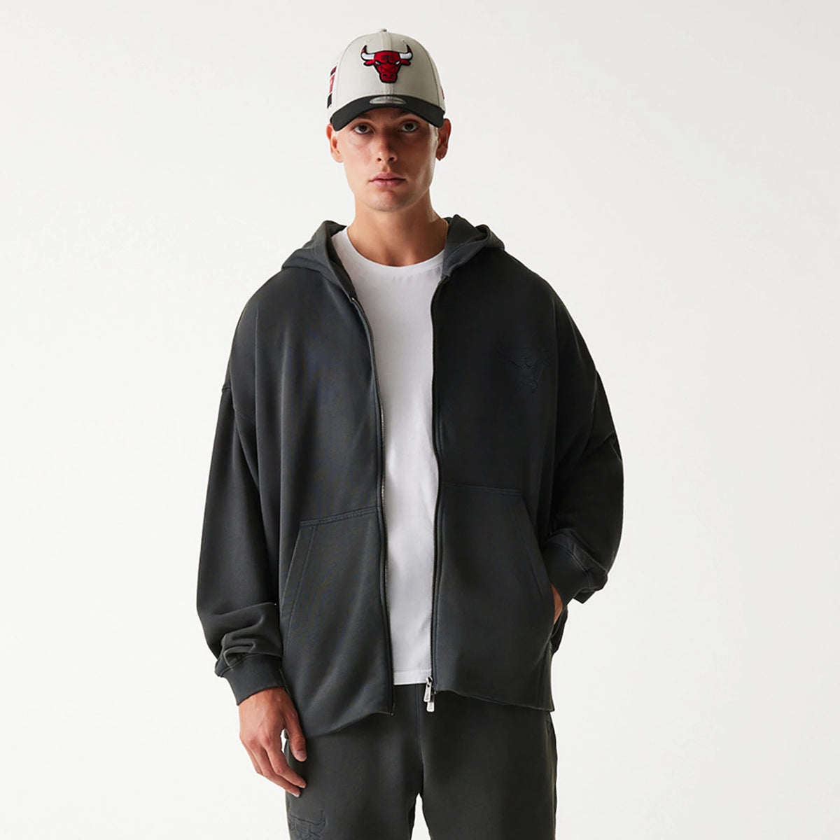 New Era Chicago Bulls NBA Washed Grey Full Zip Hoodie, on-model view showing the grey hoodie with a full front zip and a raised Bulls logo on the left chest, paired with a white cap featuring an embossed red Bulls logo.