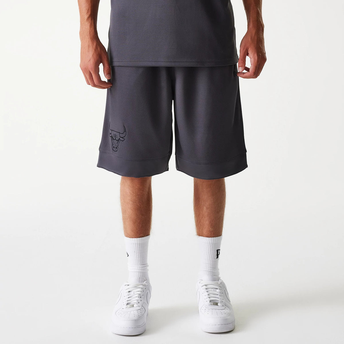 New Era Chicago Bulls NBA Tonal Dark Grey Mesh Shorts, front view showing the long dark grey shorts with an embossed Chicago Bulls logo on the left leg, loose fit ending just below the knee, paired with white socks and trainers.