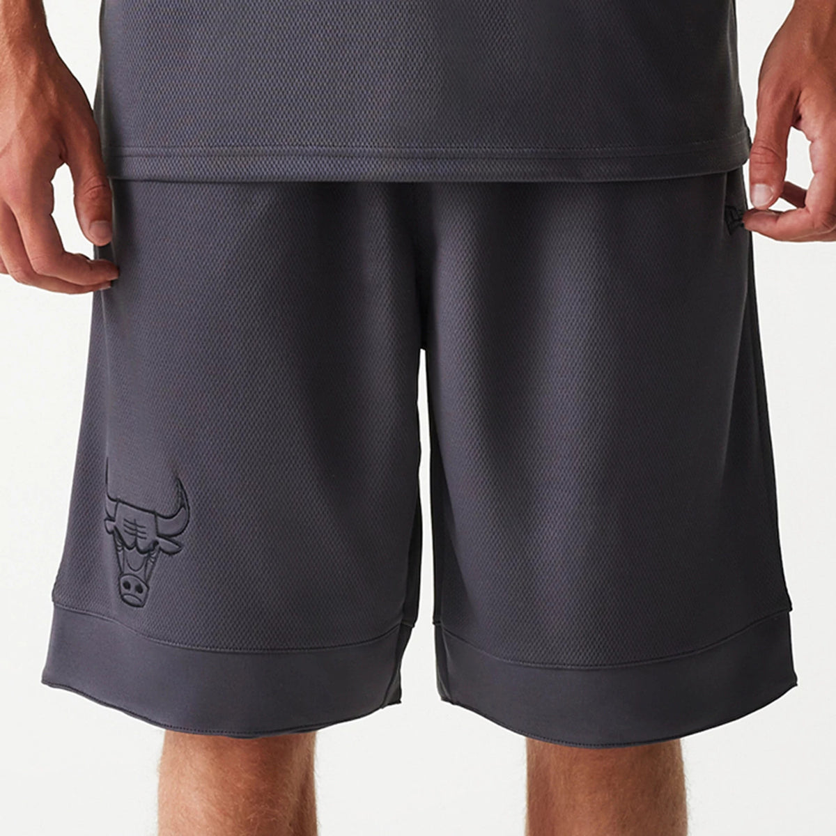 New Era Chicago Bulls NBA Tonal Dark Grey Mesh Shorts, front view showing the dark grey mesh fabric with an embossed Chicago Bulls logo on the lower left leg and a slightly longer hem at the knee.