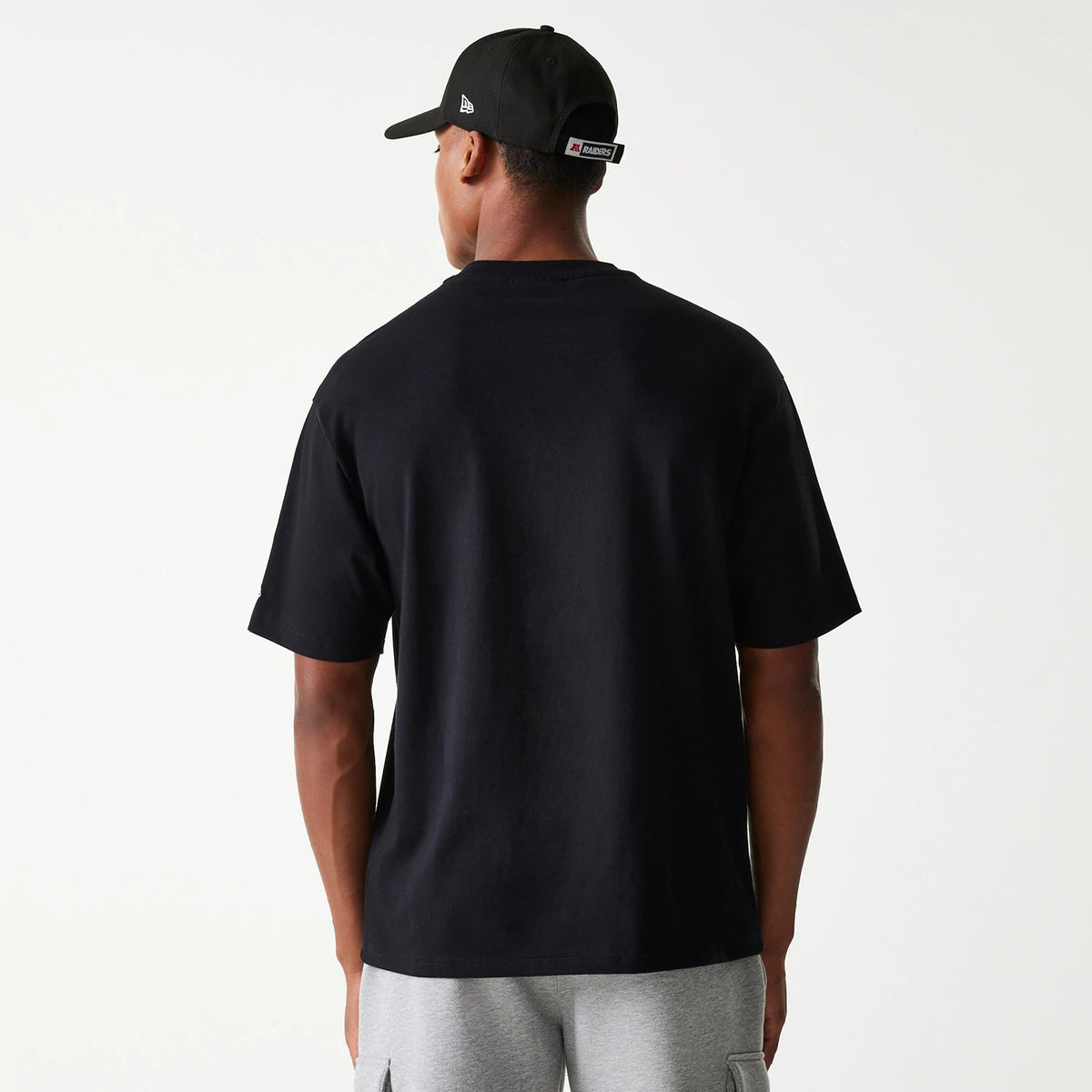 New Era Las Vegas Raiders NFL Helmet Black Oversized T-Shirt, rear view showing the plain black back with short sleeves, paired with a black cap featuring a raised logo on the side.