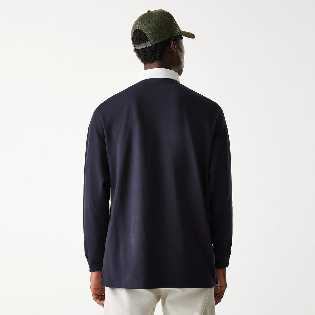 New Era Navy Oversized Rugby Shirt, back view on a model wearing a green cap, showing the relaxed fit and white collar contrasting with the navy fabric, paired with light trousers.