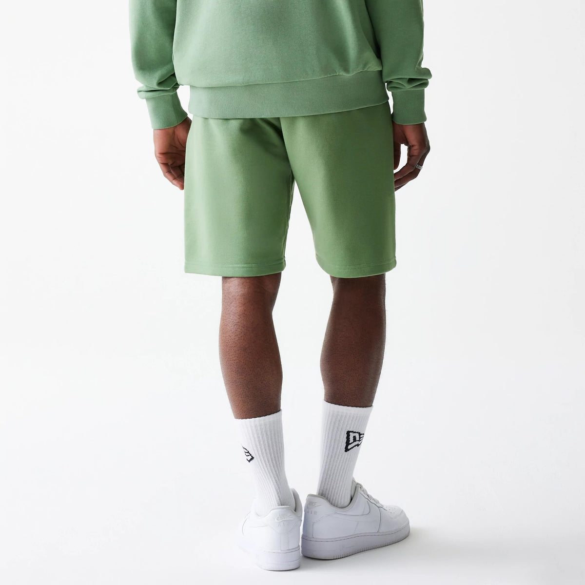 New Era Essential Green Oversized Shorts, rear view showing the relaxed fit green shorts worn by a male model, paired with a matching green top, white New Era socks featuring a raised logo, and white sneakers.