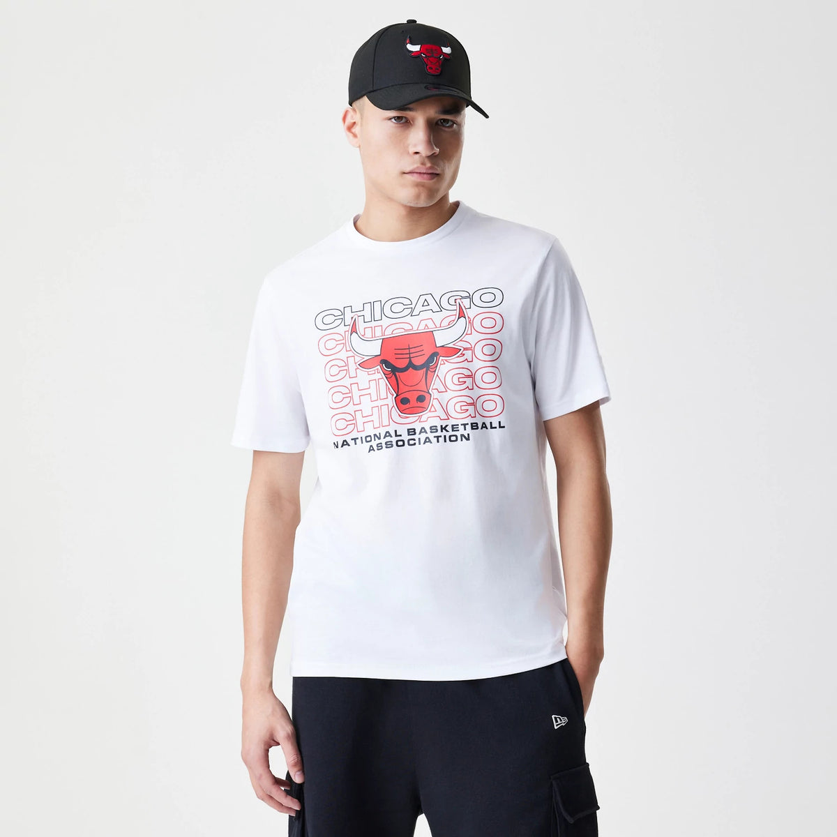 New Era Chicago Bulls NBA Stack Wordmark White T-Shirt, on-model view showing a red Bulls logo and stacked text design on the front, paired with a black cap featuring the same raised Bulls logo and plain black shorts with a small New Era logo.