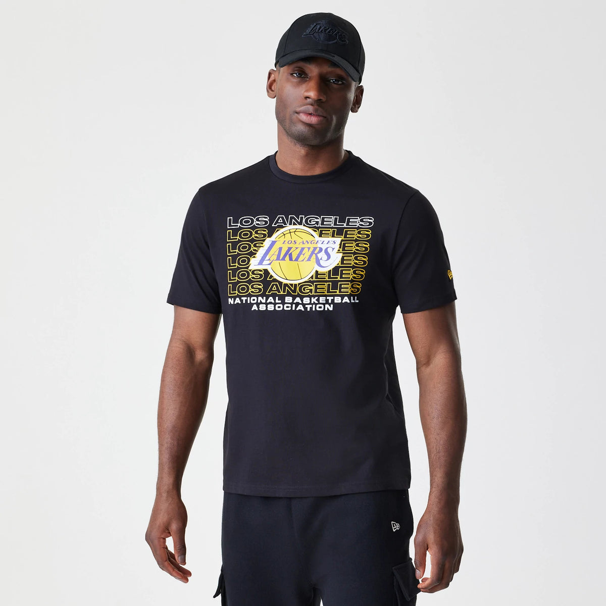 New Era LA Lakers NBA Stack Wordmark Black T-Shirt, on-model view showing the bold yellow and white LA Lakers graphic printed on the front, with a small raised New Era logo on the left sleeve and the model wearing a black cap.