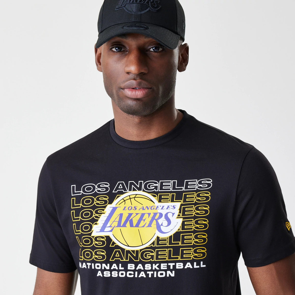 New Era LA Lakers NBA Stack Wordmark Black T-Shirt, on-model view showing the chest design with overlapping Los Angeles text in white and yellow against a basketball graphic, and a small logo on the left sleeve.