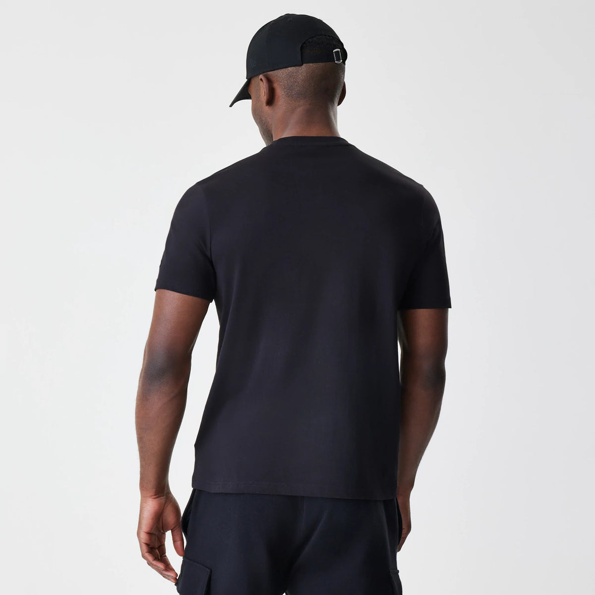New Era LA Lakers NBA Stack Wordmark Black T-Shirt, rear view showing the plain black back of the T-shirt with short sleeves, worn by a person also wearing a black cap with an adjustable strap.