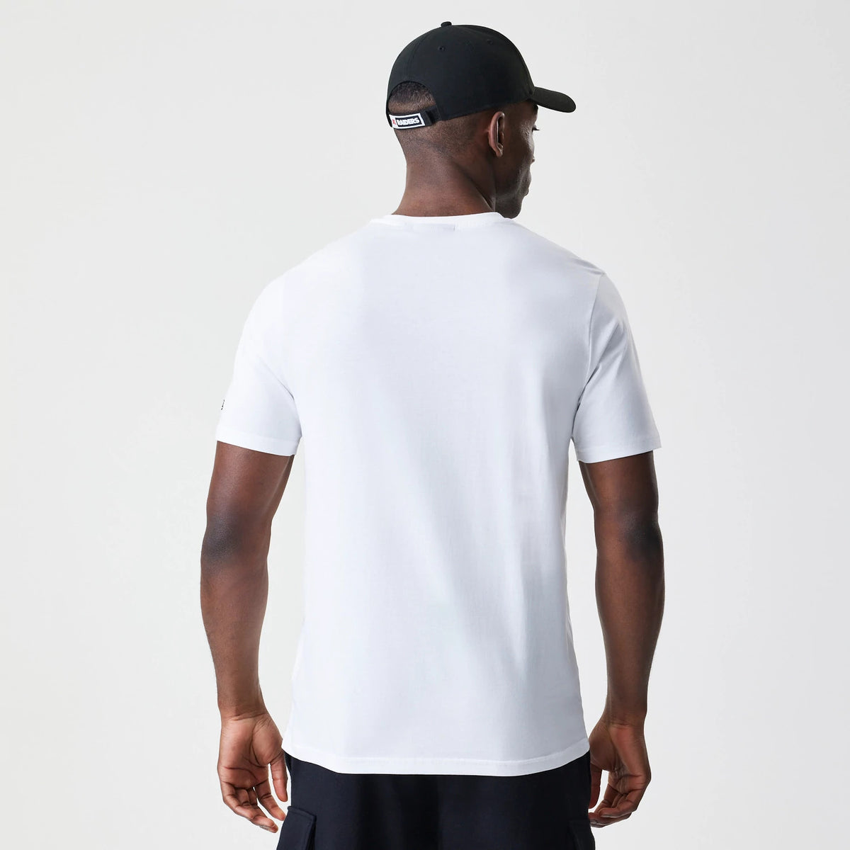 New Era Las Vegas Raiders NFL Stack Arch Logo White T-Shirt, on-model rear view showing the plain white back of the T-shirt with short sleeves, worn with a black cap featuring an embroidered patch above the adjustable strap.