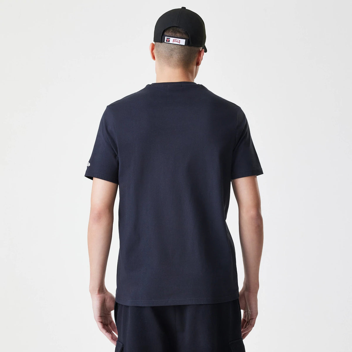 New Era Chicago Bulls NBA Logo Black T-Shirt, rear view showing a plain black back with short sleeves, a debossed New Era logo on the left sleeve, worn by a person also in a black cap featuring a debossed Chicago Bulls logo above the strap.